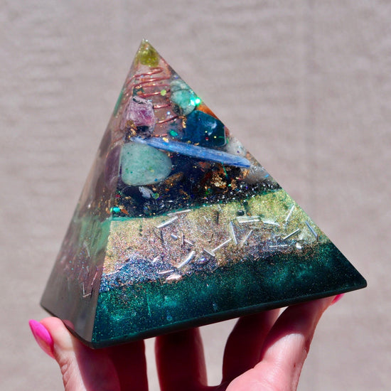 Pyramid-shaped orgonite resin crystal with embedded stones held by a hand against a neutral background