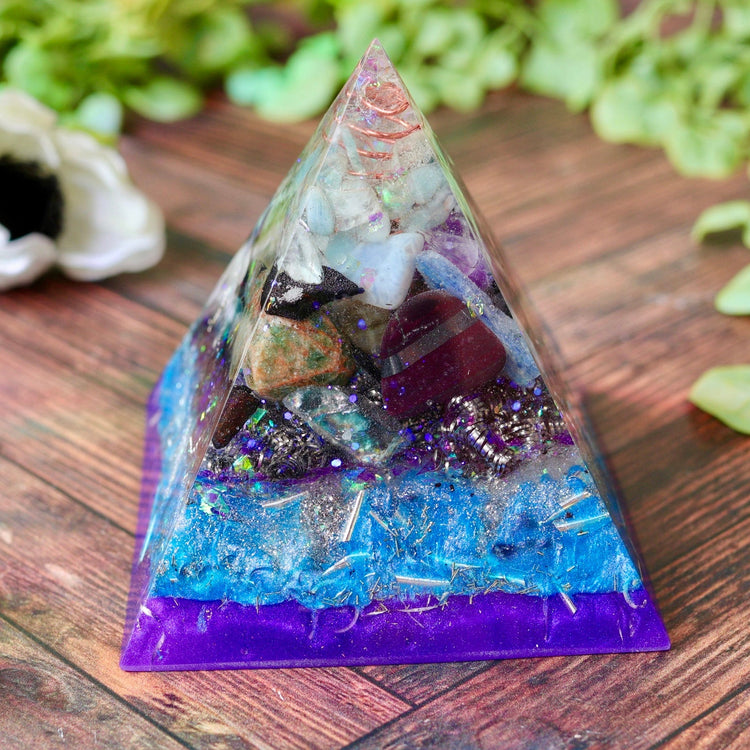 Pyramid-shaped crystal Orgonite with embedded stones on a wooden surface with greenery